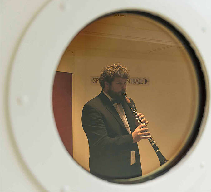 Blick durch rundes Türfenster auf einen Mann, der Klarinette spielt.
